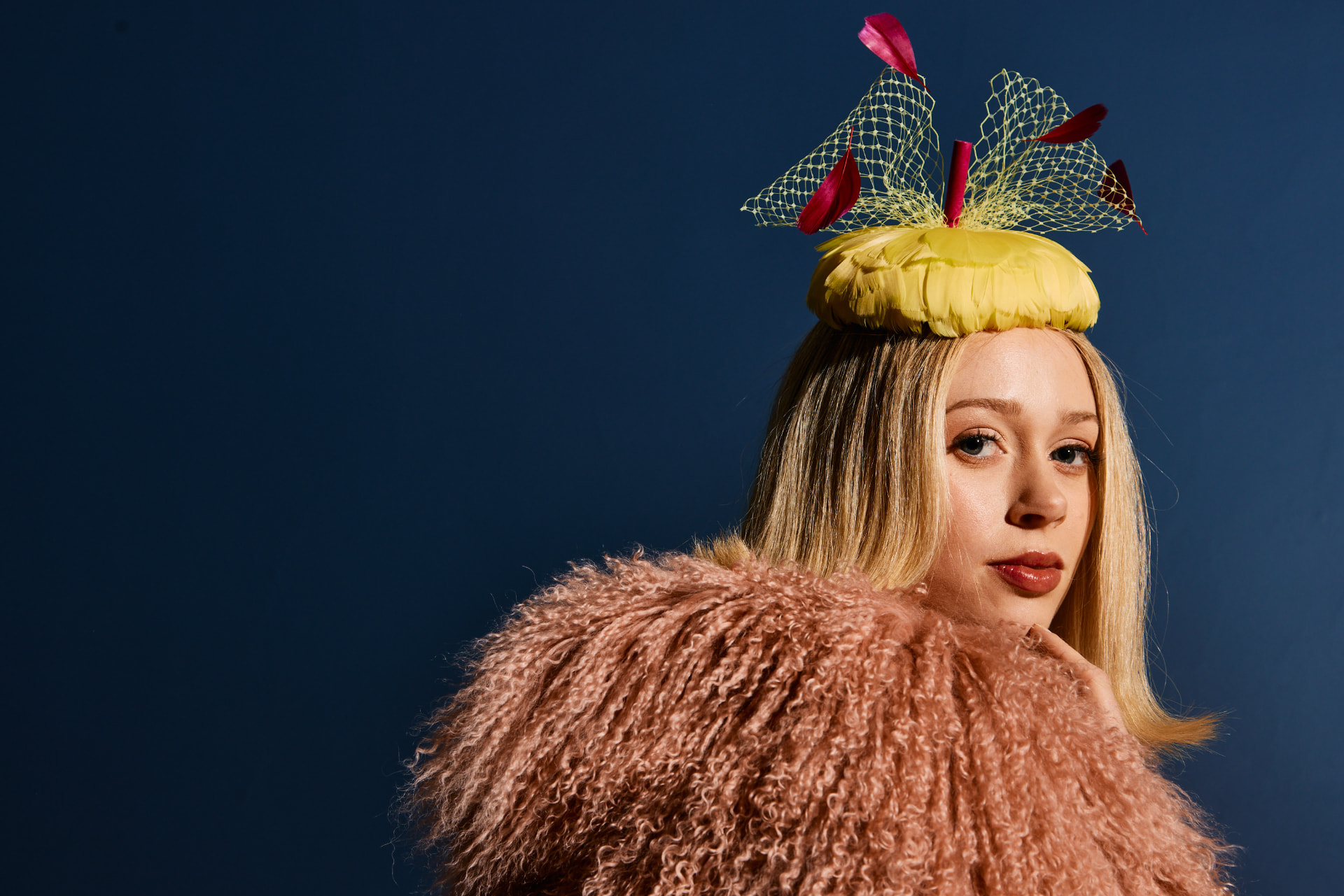 Model in a pink shearling coat wearing a yellow feathered millinery piece, against a deep navy backdrop